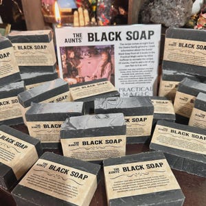 May include: Black soap bars with labels reading "The Aunts BLACK SOAP" are arranged around a sign. The sign says "BLACK SOAP" and "PRACTICAL MAGIC". The soap bars are dark gray with a light gray top. The labels list ingredients.