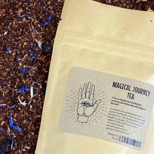 May include: A brown paper bag with a label that reads "Magical Journey Tea". The label features a hand with an eye in the palm and the text "Life is a magical journey filled with wondrous discoveries along with bumps in the road." The tea is described as a blend of blueberries, maple syrup, and honeybush tea. It is caffeine-free and contains no artificial ingredients. The bag is priced at $8.00.