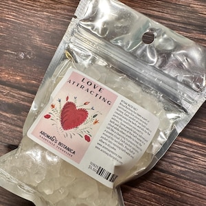 May include: A clear plastic bag filled with bath salts. The label reads "Love Attracting" with a red heart design. The brand name "Aromag's Botanica" is also visible. The bag is on a wooden surface.