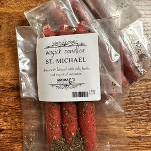 May include: Red magick candles, dressed and blessed with oils, herbs, and mystical intention. The label reads "Magick Candles, ST. MICHAEL" and includes the brand name "AROMAG'S BOTANICA". The candles are packaged in clear plastic.