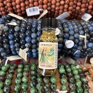 May include: A small glass bottle of Imbolc oil with a black cap, featuring a label with a woman in a green dress. The bottle is surrounded by beaded bracelets in shades of brown, blue, and green.