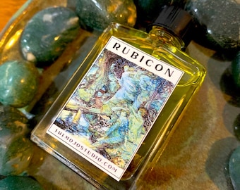 May include: A small glass bottle with a yellow liquid and a label that reads "RUBICON" and "THE MOJO STUDIO.COM". The bottle is sitting on a surface with green stones.