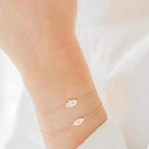 May include: Two gold bracelets with charms. One charm is a hamsa hand with a small diamond in the centre. The other charm is an evil eye with a small diamond in the centre.