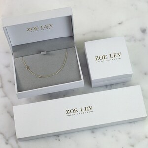 K&ouml;nnte beinhalten: Wei&szlig;e Schmuckschatulle mit goldenem Logo, auf dem "ZOE LEV - LOS ANGELES" steht. Die Schachtel ist ge&ouml;ffnet und eine goldene Kettenkette mit einem Buchstabenanh&auml;nger befindet sich darin.