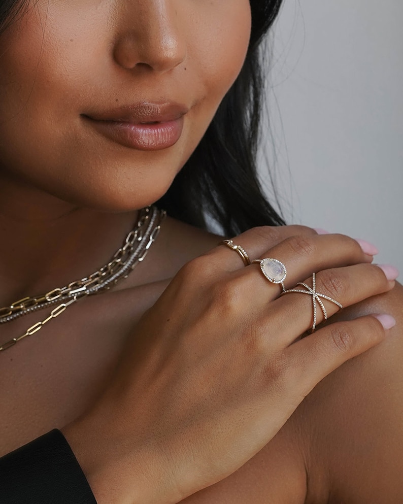K&ouml;nnte beinhalten: Ein goldener Ring mit einem birnenf&ouml;rmigen wei&szlig;en Edelstein und einem Kreuz-Design. Der Ring wird an einem Finger getragen.