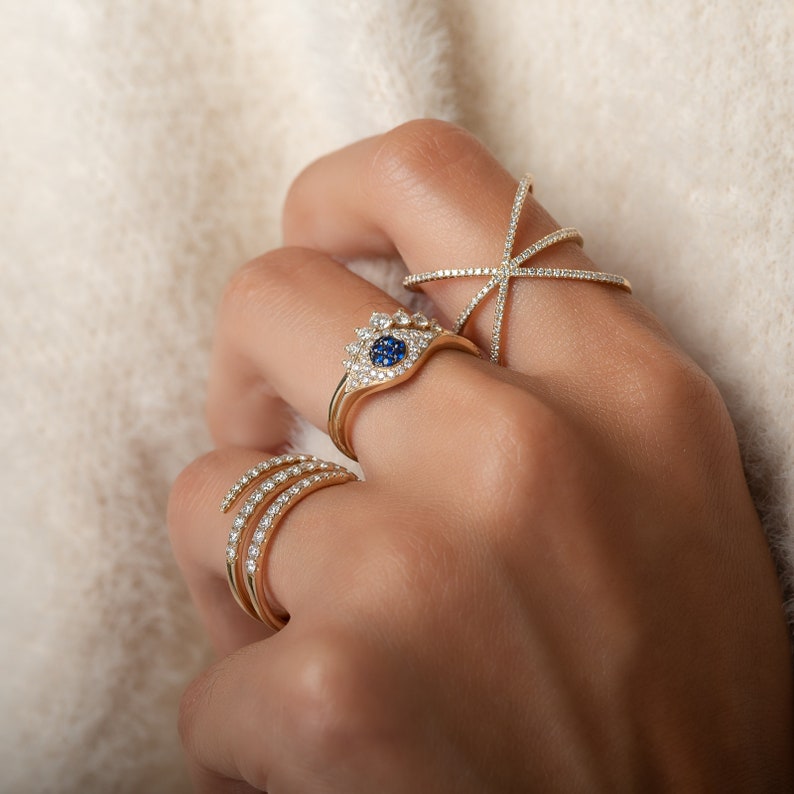 May include: A hand wearing four gold rings. One ring features a blue gemstone surrounded by smaller white stones. Another is a crisscross design, and a third has three rows of white stones. The fourth ring is a simple band.