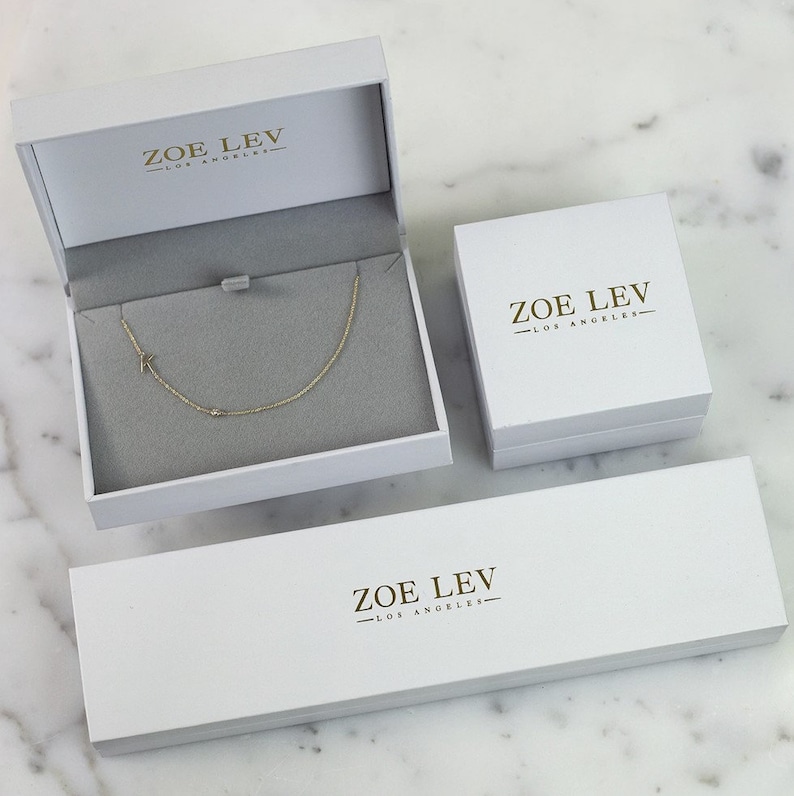 May include: A gold necklace with the letter "K" in an open white jewelry box. Two additional white boxes are also present, all with the text "ZOE LEV - LOS ANGELES" in gold. The necklace is displayed on a gray interior.