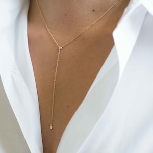 May include: Delicate gold-tone necklace with a Y-shaped design. The necklace features three small, round, clear gemstones. The chain is thin and hangs down the chest. The necklace is worn over a white shirt.