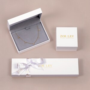 May include: White gift box with a gold ribbon and the text "ZOE LEV - LOS ANGELES" on the lid. Inside the box is a gold necklace with the letters "SOFI" on it.