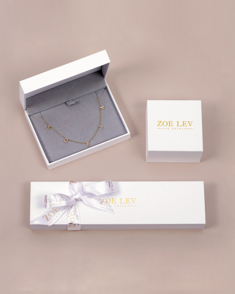 May include: White gift box with a gold ribbon and the text "ZOE LEV - LOS ANGELES" on the lid. Inside the box is a gold necklace with the letters "SOFI" on it.