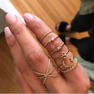 May include: A hand displaying a collection of gold rings. The rings feature various designs, including a band with small stones, a ring with a small opal, a ring with a baguette-cut stone, and rings with the letters "S", a crescent moon, and a star.