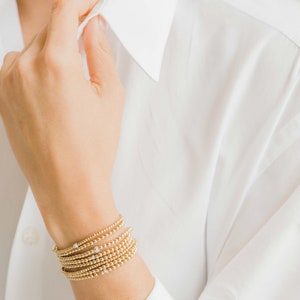 Pode incluir: Um conjunto de pulseiras de contas douradas com pequenas pedras transparentes sobre um fundo branco.