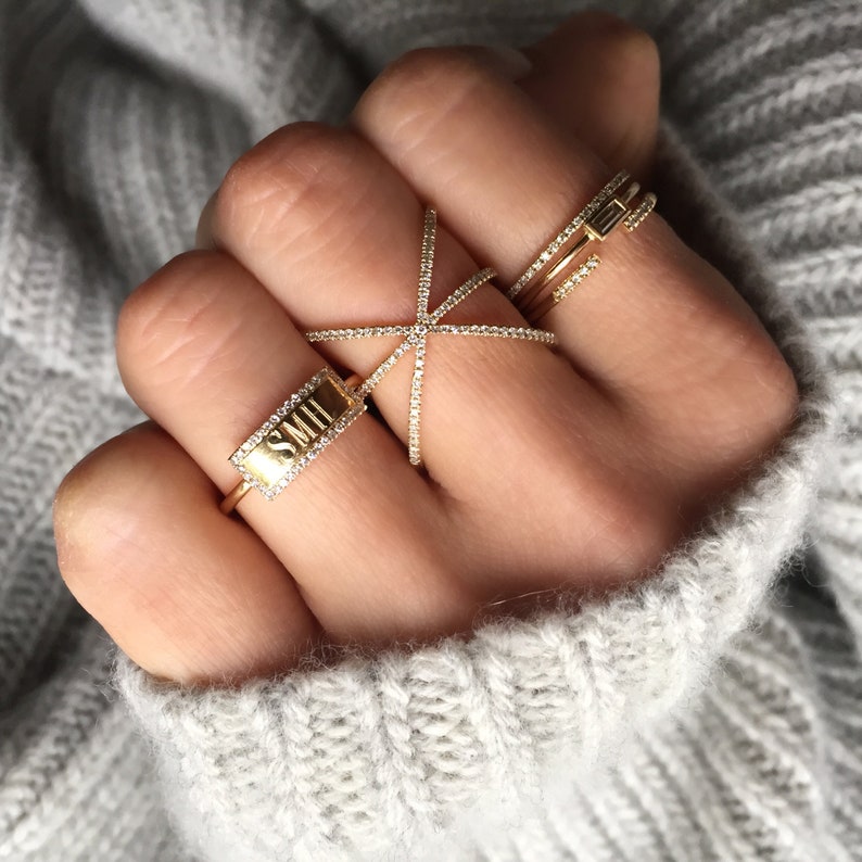 May include: A hand wearing several gold rings. One ring has the initials "SMII" with a diamond border. Another ring has a starburst design. A third ring has a rectangular diamond. The rings are on a hand with a gray sweater in the background.