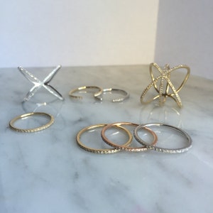 K&ouml;nnte beinhalten: Ein Set aus f&uuml;nf Diamantringen in verschiedenen Stilen. Die Ringe sind aus Gold und Silber und haben verschiedene Designs, darunter ein schlichtes Band, ein Kreuzdesign und ein filigraneres Design mit mehreren B&auml;ndern.