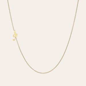 以下が含まれることがあります： 繊細なゴールドカラーのネックレスで、筆記体の「A」のペンダントが特徴です。ペンダントは細いチェーンに取り付けられており、ミニマルでエレガントなデザインになっています。ネックレスはニュートラルな背景に表示されています。