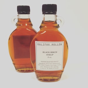 May include: Two clear glass bottles of Halifax Hollow Black Birch Syrup, each with a black cap and a small handle. The labels read "Black Birch Syrup 8 oz." and list ingredients. The syrup is amber-colored.