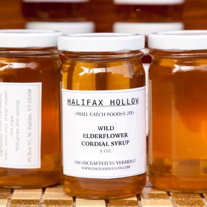 May include: Three glass jars filled with a golden-colored syrup. Each jar has a white lid and a white label with black text. The labels read "Wild Elderflower Cordial Syrup" and "Handcrafted in Vermont".