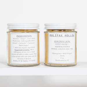 Puede incluir: Dos frascos de vidrio con tapas y etiquetas blancas. Una etiqueta enumera ingredientes como c&uacute;rcuma y jengibre. La otra etiqueta dice "Halifax Hollow Golden Lion: Lion's Mane + Ashwagandha Herbal Golden Milk Mix". Los frascos est&aacute;n llenos de un polvo dorado.