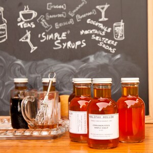 May include: Bottles of Halifax Hollow cinnamon stick simple syrup are displayed on a wooden surface. A clear glass mug and a whisk sit on a silver tray. A chalkboard in the background features the words "Simple Syrups" and drawings of cocktails and teas.