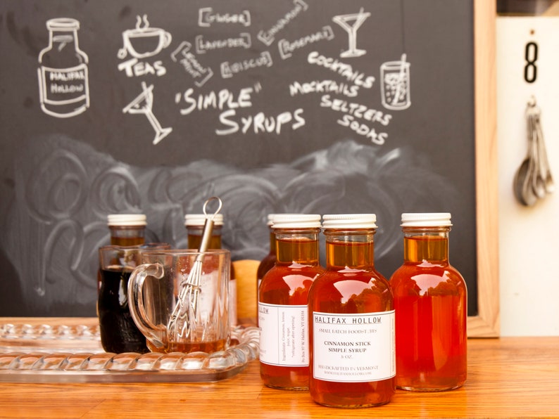 May include: Bottles of Halifax Hollow cinnamon stick simple syrup are displayed on a wooden surface. A clear glass mug and a whisk sit on a silver tray. A chalkboard in the background features the words "Simple Syrups" and drawings of cocktails and teas.