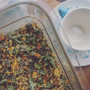 May include: A clear rectangular container filled with dried herbs and flowers, including yellow, orange, and green elements. A white teacup and saucer with a blue handle and rim design are in the background.