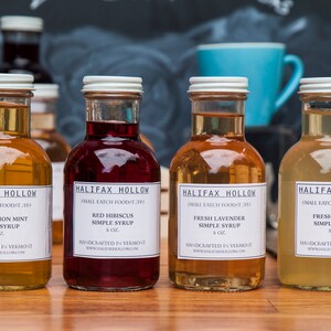May include: Four glass bottles of simple syrup with white labels. Flavors include Fresh Lemon Mint, Red Hibiscus, Fresh Lavender, and Fresh Ginger. Each bottle is 8 oz and handcrafted in Vermont. The bottles are arranged on a wooden surface.