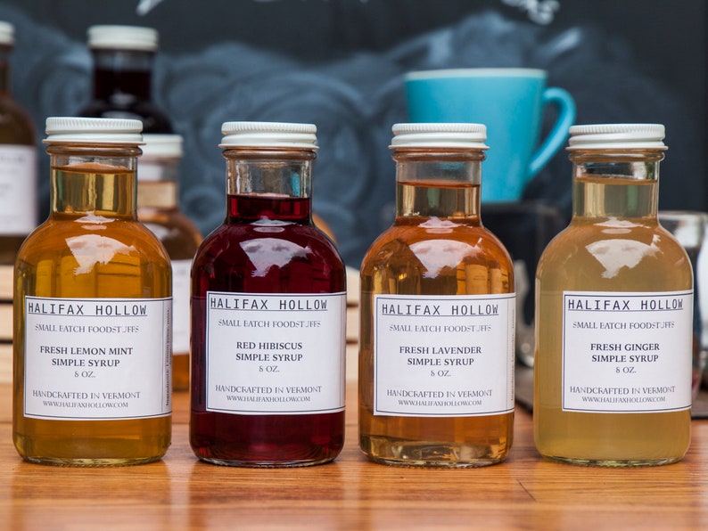 May include: Four glass bottles of simple syrup with white labels. Flavors include Fresh Lemon Mint, Red Hibiscus, Fresh Lavender, and Fresh Ginger. Each bottle is 8 oz and handcrafted in Vermont. The bottles are arranged on a wooden surface.