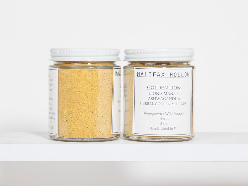 Puede incluir: Dos frascos de vidrio con tapas blancas llenos de un polvo dorado. Los frascos tienen etiquetas que dicen "HALIFAX HOLLOW" y "GOLDEN LION: LION'S MANE + ASHWAGANDHA HERBAL GOLDEN MILK MIX". Los frascos est&aacute;n sobre una superficie blanca.