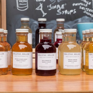 May include: A collection of glass bottles filled with colorful syrups, labeled "Halifax Hollow" and various flavors like lavender, lemon mint, and ginger. The bottles are arranged on a wooden surface, with a chalkboard background displaying "Simple Syrups."