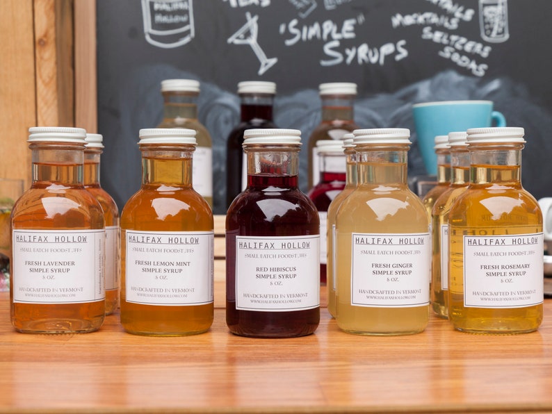 May include: A collection of glass bottles filled with colorful syrups, labeled "Halifax Hollow" and various flavors like lavender, lemon mint, and ginger. The bottles are arranged on a wooden surface, with a chalkboard background displaying "Simple Syrups."