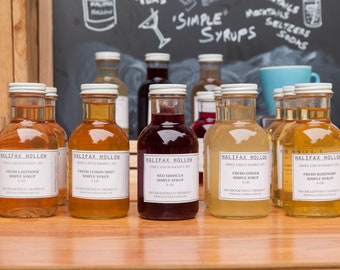 Puede incluir: Una colección de botellas de vidrio llenas de coloridos siropes, etiquetadas como "Halifax Hollow" y con varios sabores como lavanda, menta de limón y jengibre. Las botellas están dispuestas sobre una superficie de madera, con un fondo de pizarra que muestra "Simple Syrups."