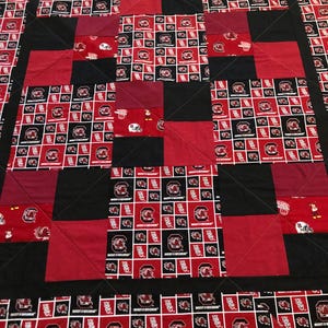 May include: A handmade quilt featuring a repeating pattern of red, black, and white squares. The squares display the University of South Carolina Gamecocks logo and football helmets. The quilt is bordered with black fabric and has a quilted design.
