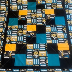 May include: A quilt made of fabric squares featuring the Jacksonville Jaguars logo and the words "Jaguars" and "JAGS". The quilt is made of black, gold, and teal fabric.
