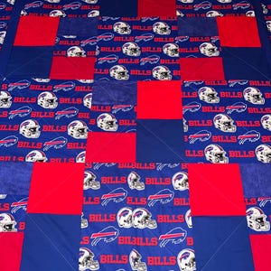 May include: A blue and red patchwork quilt with a Buffalo Bills theme. The quilt is made of squares with the Bills logo and the words "Bills" repeated on each square.