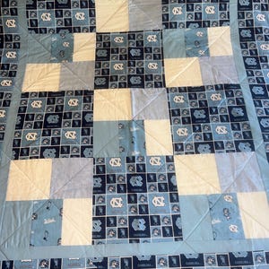 May include: A patchwork quilt in shades of blue and white. The quilt features squares of solid white, light blue, and patterned fabric with the word "CAROLINA" and a logo. The quilt is likely handmade.
