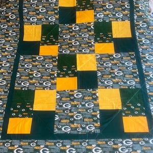 May include: A green and gold patchwork quilt with a repeating pattern of the Green Bay Packers logo. The quilt is made up of squares of different sizes and colors.