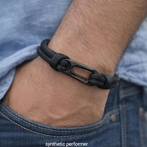 May include: A black cord bracelet featuring a matte black metal carabiner-style clasp. The bracelet is constructed from two parallel strands of woven cord that loop through the metal hardware, creating a secure and functional closure.
