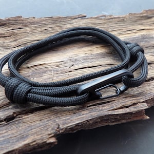 May include: A black paracord bracelet with a black metal clasp. The bracelet is made of thick, woven rope and is looped around itself. The background is a piece of weathered wood.