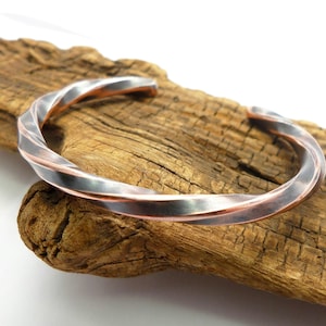 May include: A cuff bracelet featuring a twisted design made from two distinct metal strands. One strand has a polished silver-toned finish, while the other strand displays a warm, metallic copper-toned finish, creating a contrasting two-tone effect.