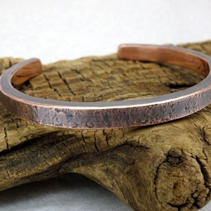 May include: A copper bracelet with a hammered finish. The bracelet is a simple, minimalist design and has a slightly rounded shape.