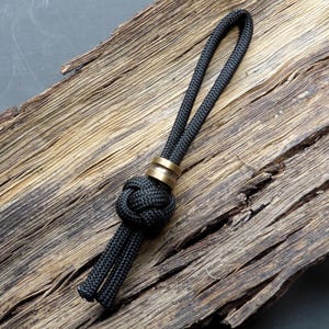 May include: A black rope keychain with a decorative knot and a brass-colored metal ring. The keychain is displayed on a piece of weathered wood with a dark background. The rope is approximately 10 inches long.