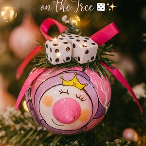 Puede incluir: Un adorno navide&ntilde;o redondo con un cerdo pintado que lleva una corona amarilla, rematado con dos dados blancos y una cinta rosa brillante. El texto "The Luckiest Ornament on the Tree" aparece arriba con un icono de dado.