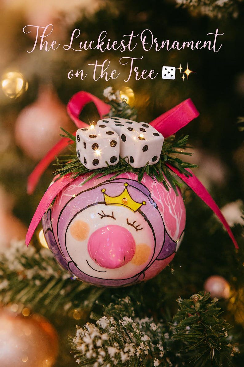 Puede incluir: Un adorno navide&ntilde;o redondo con un cerdo pintado que lleva una corona amarilla, rematado con dos dados blancos y una cinta rosa brillante. El texto "The Luckiest Ornament on the Tree" aparece arriba con un icono de dado.