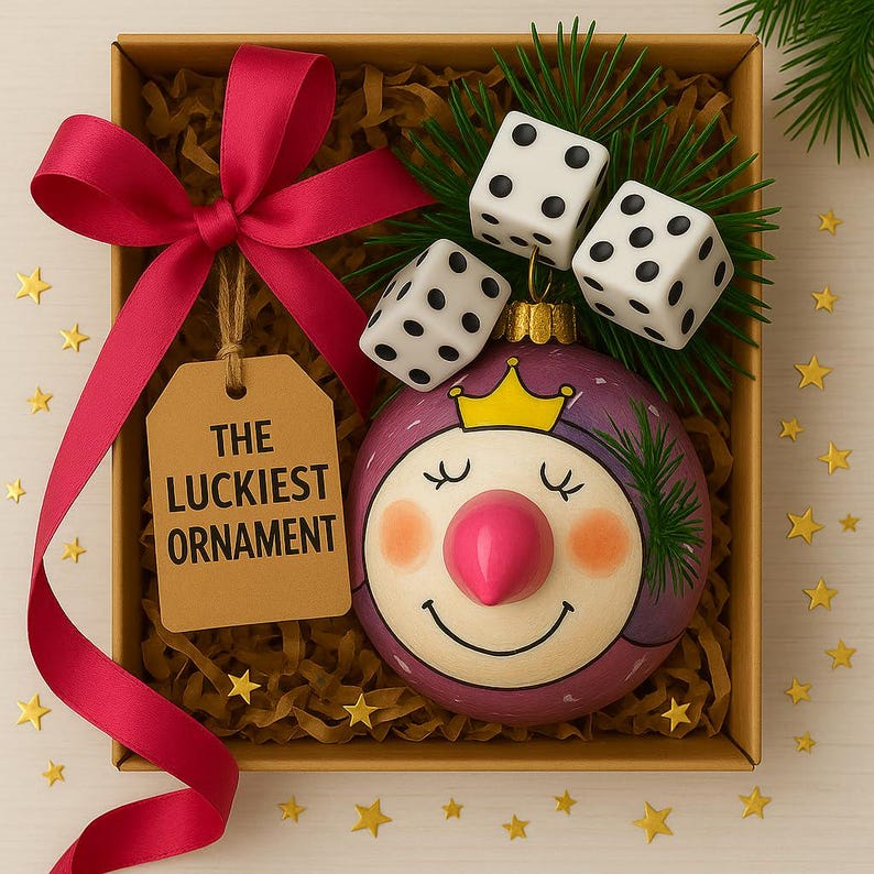 Puede incluir: Un adorno redondo de color morado con una cara pintada y una corona amarilla, decorado con tres dados blancos de puntos negros y agujas de pino verdes. Una etiqueta marr&oacute;n dice THE LUCKIEST ORNAMENT, atada con una cinta rosa vibrante.