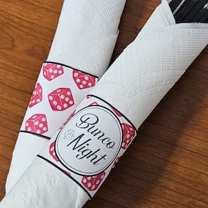 May include: Two white paper napkins rolled up, each with a decorative band. One band features red dice on a white background. The other band has the text "Bunco Night" in black and red, with a dice graphic.