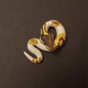 May include: A handcrafted, coiled snake figurine. The snake is primarily white with yellow and brown markings. The head is painted with brown, yellow, and black details. The figurine is set against a dark background.