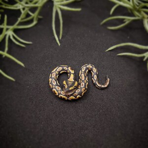 Op de afbeelding: Een handgemaakt, opgerold slangenbeeldje met een gedetailleerd patroon van zwarte, bruine en gele vlekken. De kop van de slang is omhoog en het lichaam is gebogen. Het beeldje staat tegen een donkere achtergrond, met groen gebladerte in de bovenhoeken.