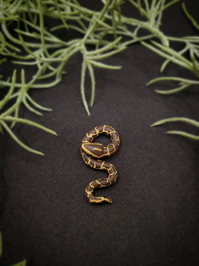 K&ouml;nnte beinhalten: Eine detaillierte, Miniatur-Schlangenskulptur mit braunem K&ouml;rper und gelben Markierungen. Die Schlange ist auf einer schwarzen Oberfl&auml;che aufgerollt, mit gr&uuml;nem Laub im Hintergrund. Die Figur ist ein dekoratives Objekt.