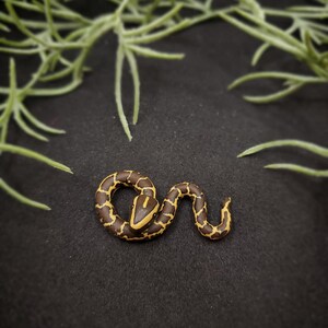 K&ouml;nnte beinhalten: Handgefertigte Miniatur-Schlangenskulptur. Die Schlange ist braun mit gelbem Muster und Kopf. Die Figur liegt auf einer schwarzen Oberfl&auml;che, mit gr&uuml;nem Laub im Hintergrund.