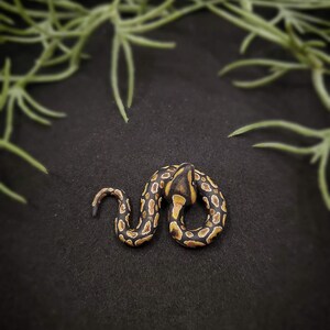 Op de afbeelding: Een handgemaakt miniatuur slangenbeeldje. De slang is opgerold, met een zwart en bruin patroon en een gele kop. Het beeldje staat tegen een donkere achtergrond, met groen gebladerte in de bovenhoeken.